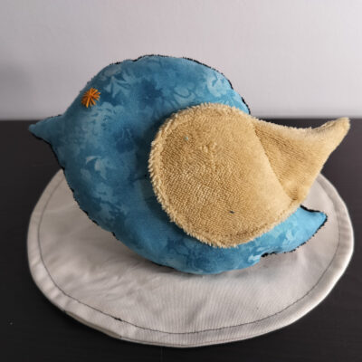 Peluche oiseau bleu en tissu marbré avec une aile en éponge jaune beige et un œil étoile orange brodé main.