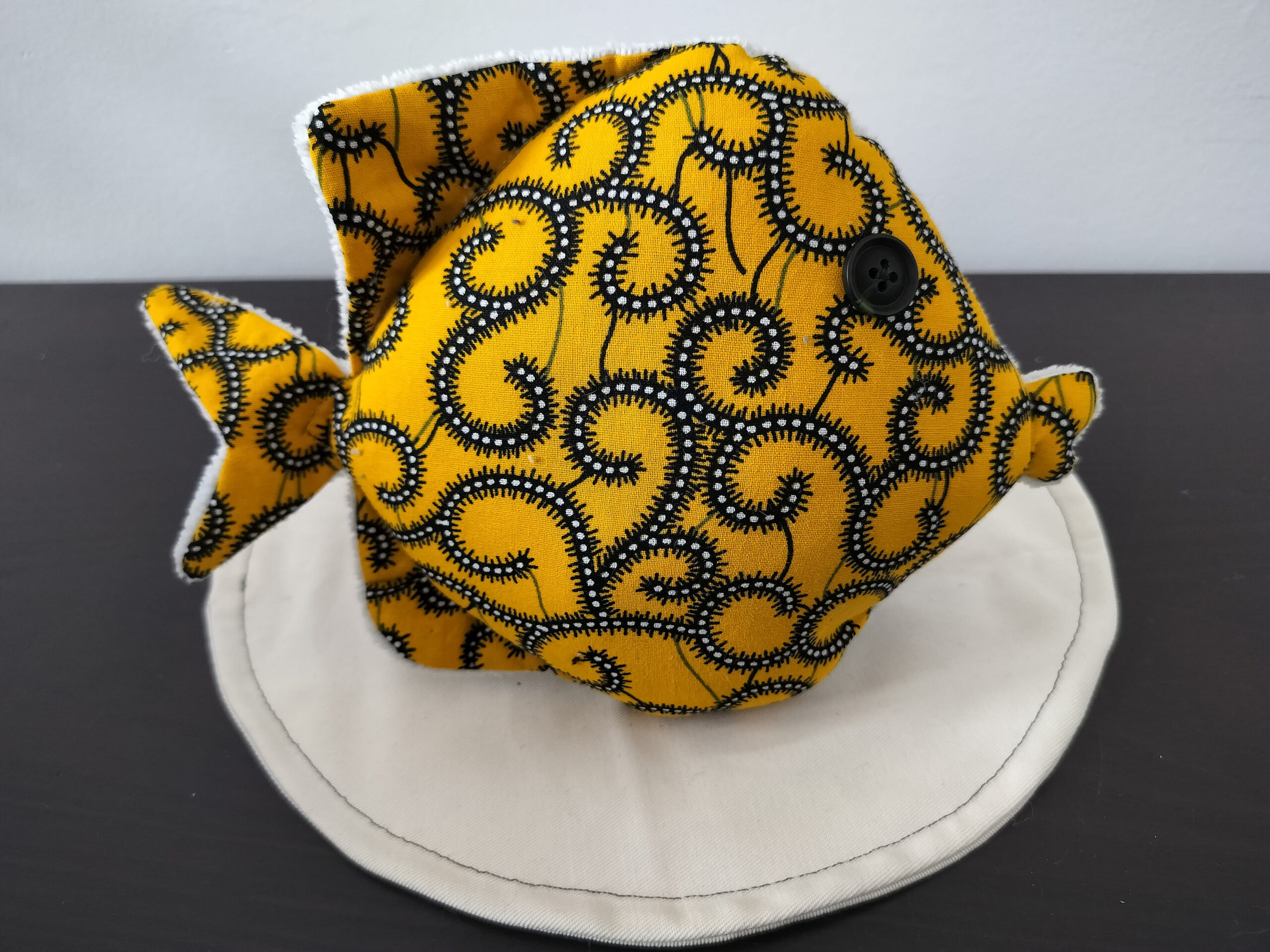 Peluche poisson rond de la collection Cameroun en tissu Wax jaune à motifs spirales avec un œil bouton noir classique.