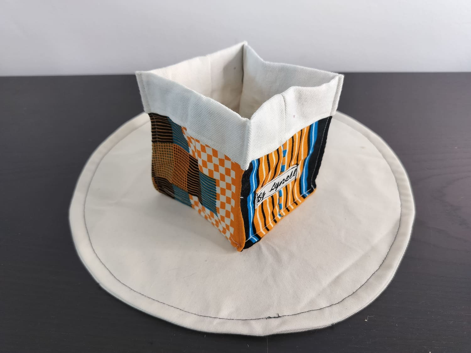 Vide-poche en tissu wax orange, petit panier carré bicolore avec revers blanc un tissu Wax aux nuances chaleureuses d'orange, de bleu et de noir, aux motifs géométriques et damiers.